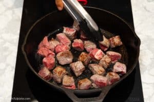 beef cubes being seared in a cast iron pan.