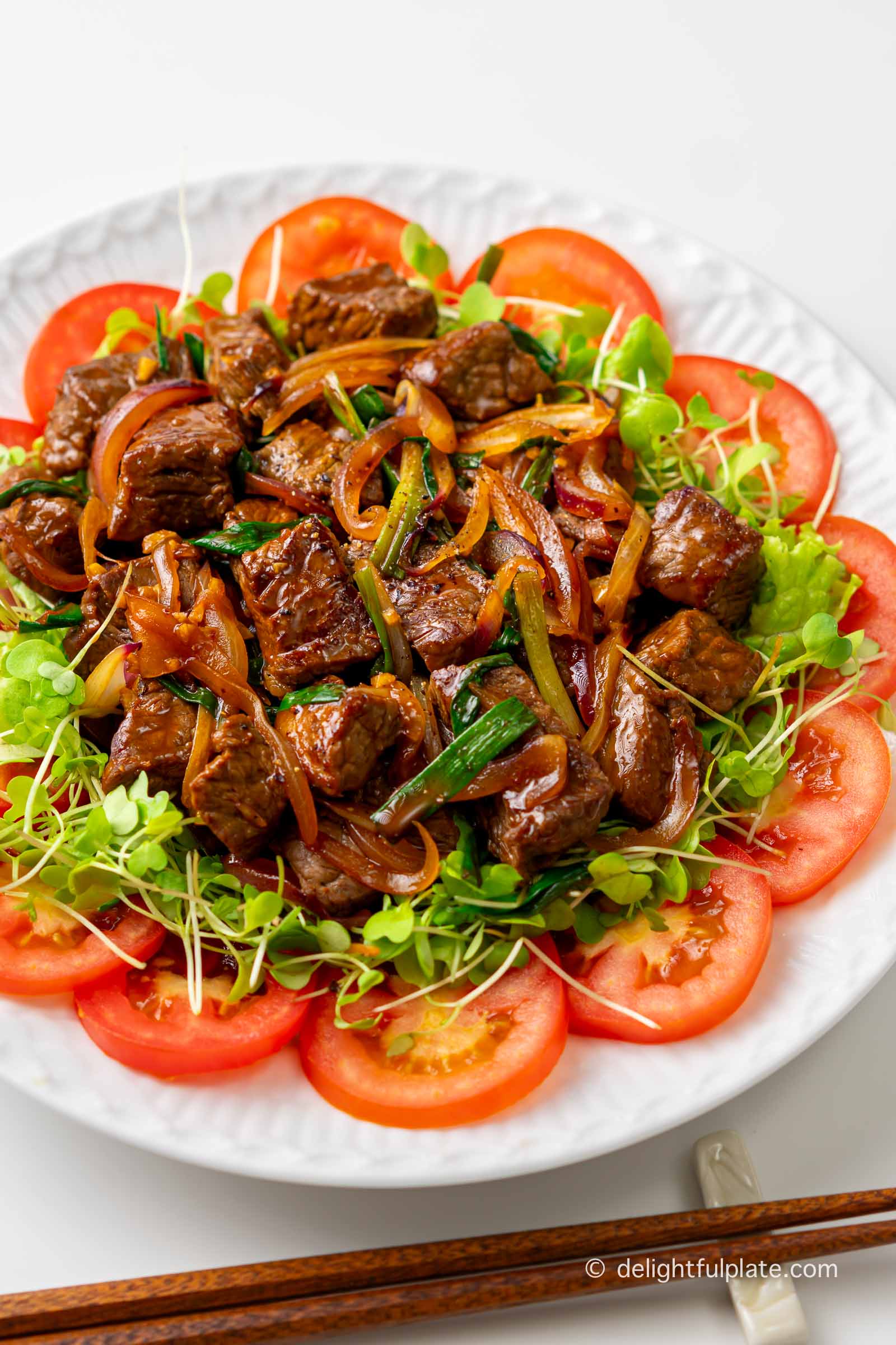 a plate of Vietnamese shaking beef (bo luc lac) placed on a bed of greens and tomatoes.
