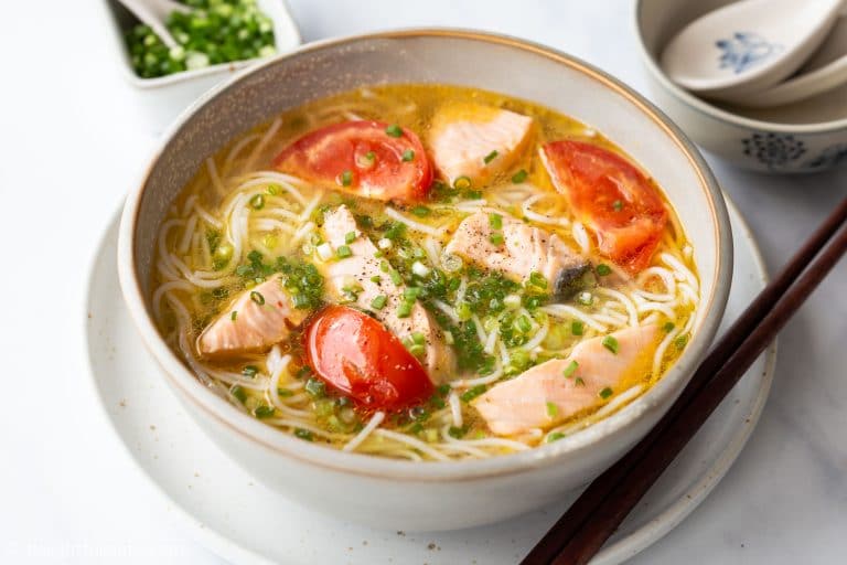 A bowl of noodle soup with pieces of salmon, tomato slices, and garnished with chopped green onions. The soup is served alongside a pair of chopsticks and a small bowl of green onions on a light-colored surface.