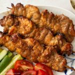 Japanese-inspired pork skewers on a serving plate with cucumber and cherry tomato